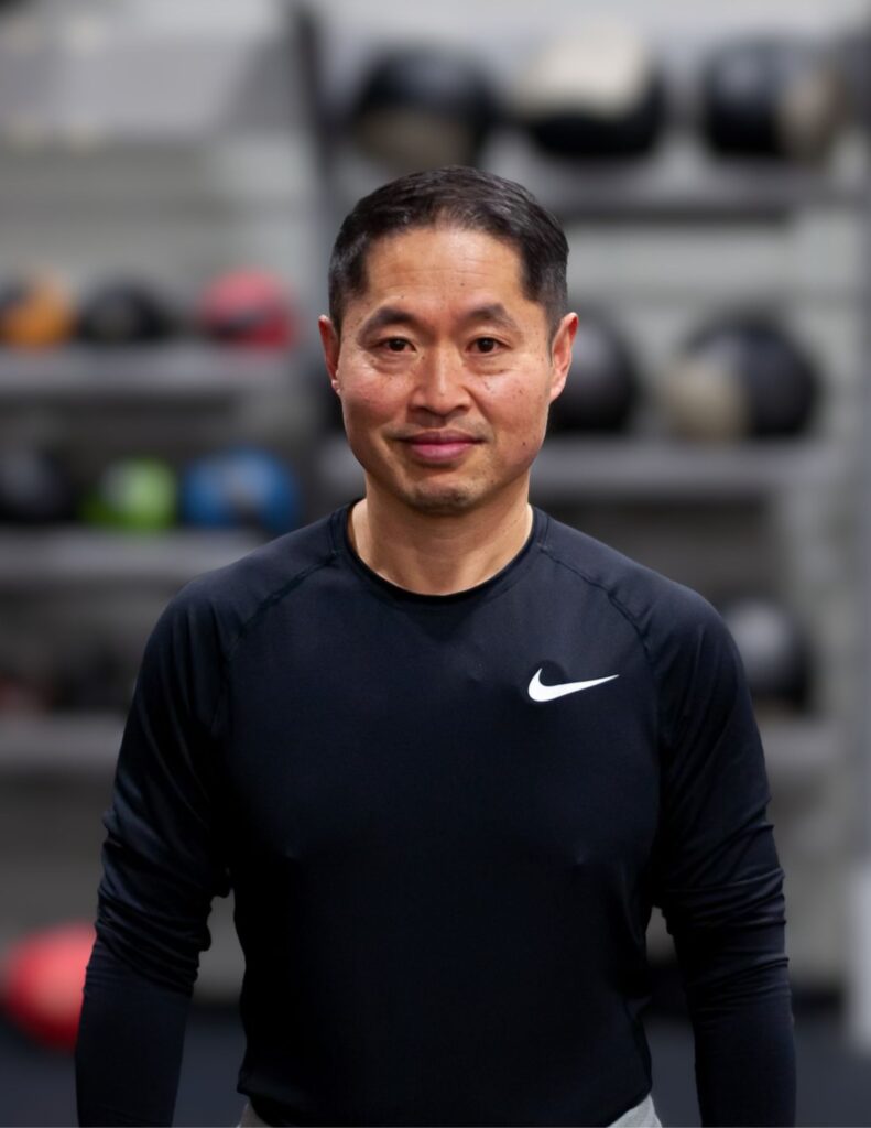 Portrait of a man in a black Nike athletic shirt standing in a gym with blurred equipment behind him.