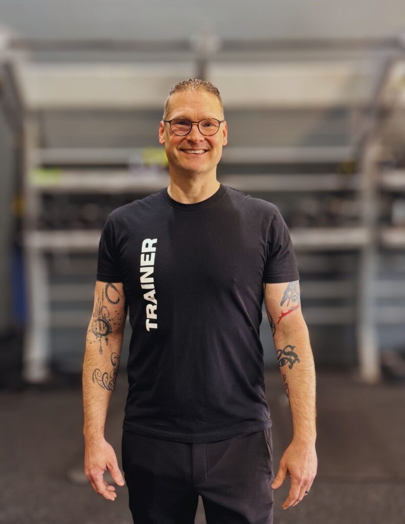 Smiling man in a black athletic shirt with tattoos on both arms, standing in a gym with blurred equipment in the background.
