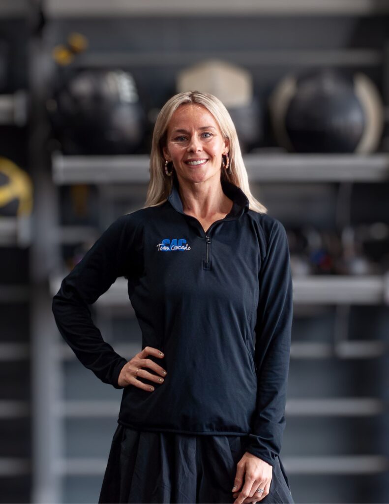 Smiling woman in a black zip-up athletic top standing in a gym with blurred exercise equipment in the background.