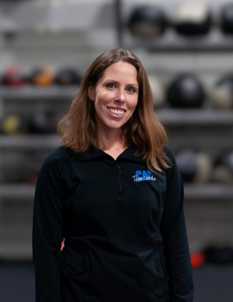 Smiling woman with shoulder-length brown hair wearing a black polo shirt, standing in a workshop or gym with blurred equipment in the background.