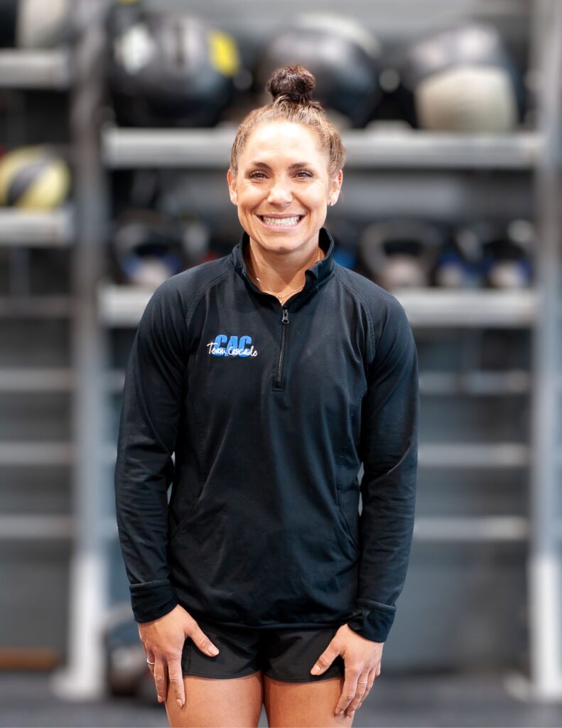 Smiling woman in a black athletic jacket standing in a gym with blurred gym equipment in the background.