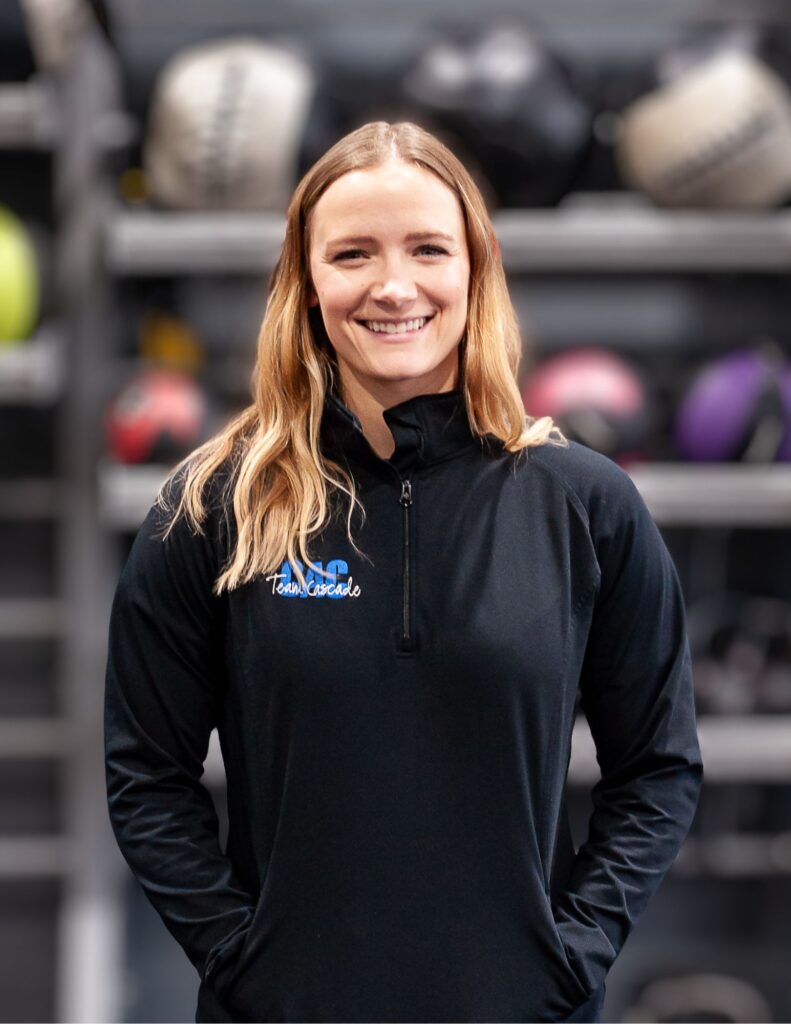 Smiling woman with long blonde hair in a black athletic jacket, gym background with blurred equipment. Jacket shows 'Team Cascade' logo.