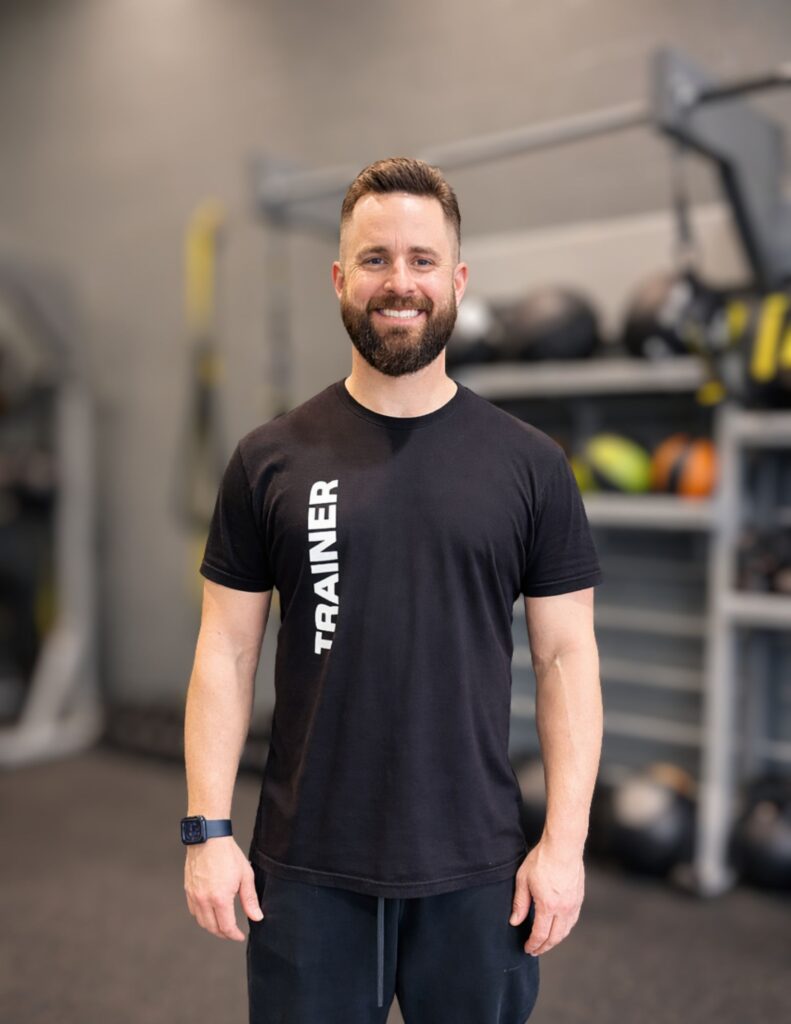 Smiling man with a beard wearing a black T-shirt that says 'TRAINER', standing in a gym with blurred equipment in the background.