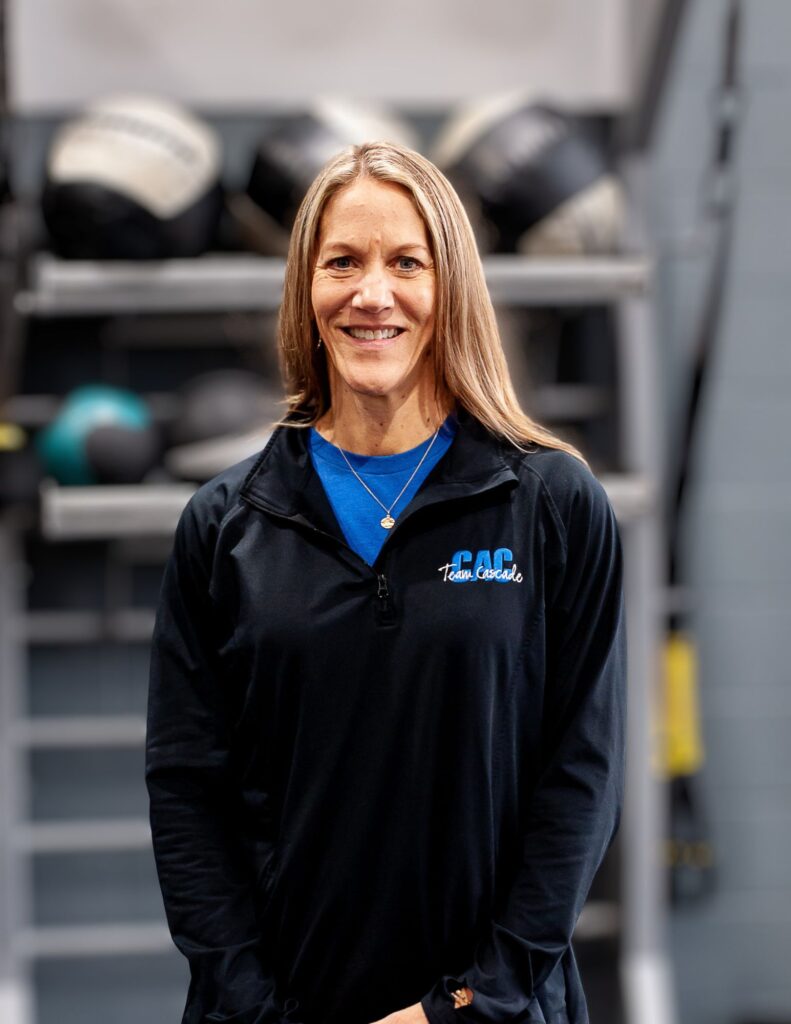 Smiling woman with long blonde hair wearing a blue shirt under a black jacket, standing in a gym with equipment blurred in the background, facing the camera.