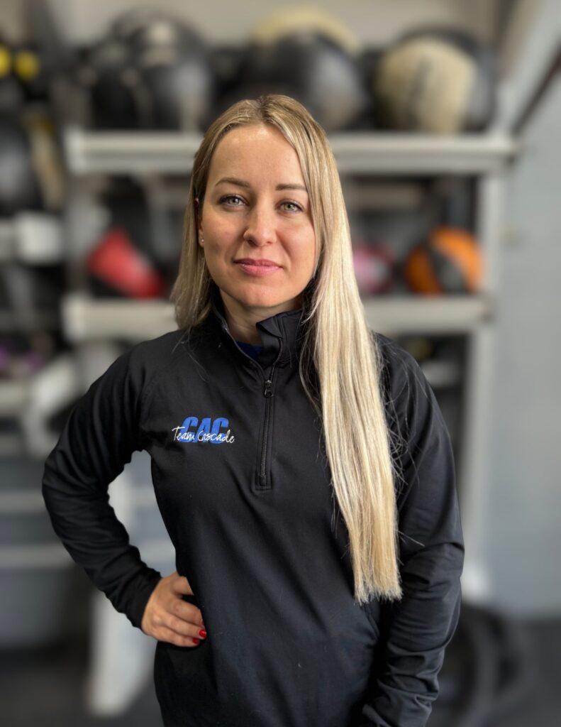 Portrait of a woman with long blonde hair in a black zip-up jacket, standing in a workshop with shelves of gear behind her.