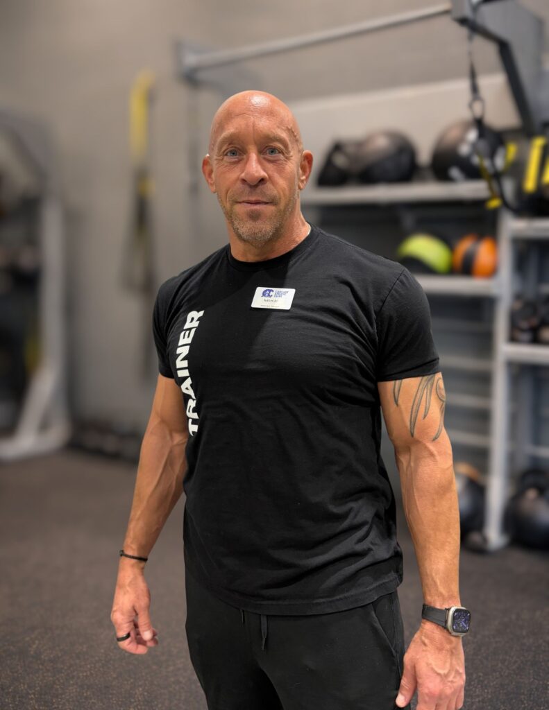 Bald man in a black athletic shirt with a name badge stands in a gym, facing the camera.