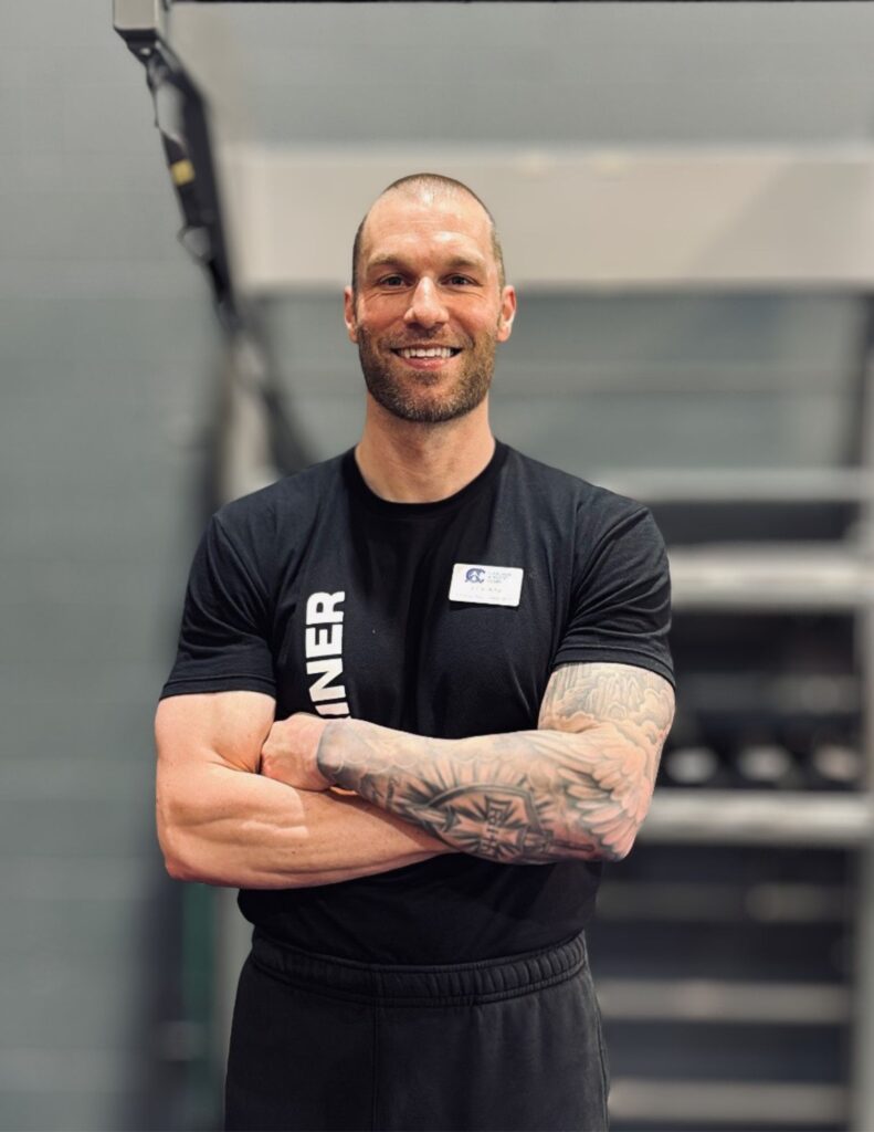 Smiling man with arms crossed in a gym, wearing a black shirt and a name badge.