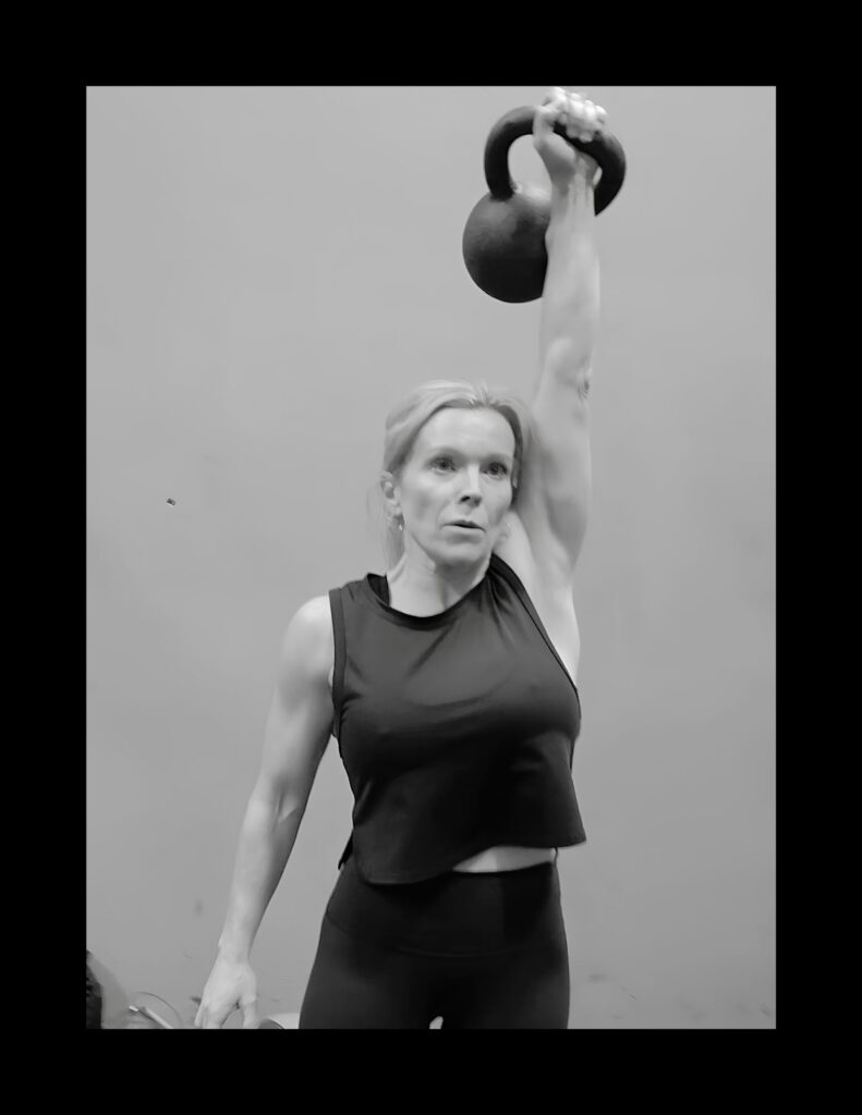 Woman in athletic wear raises a kettlebell overhead with one arm, performing a single-arm lift against a plain background (black-and-white image).