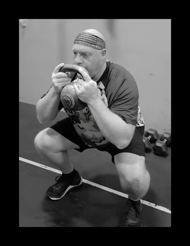 Man in a gym performing a goblet squat with a kettlebell held at chest level, wearing a striped headband and athletic clothing (BW photo).