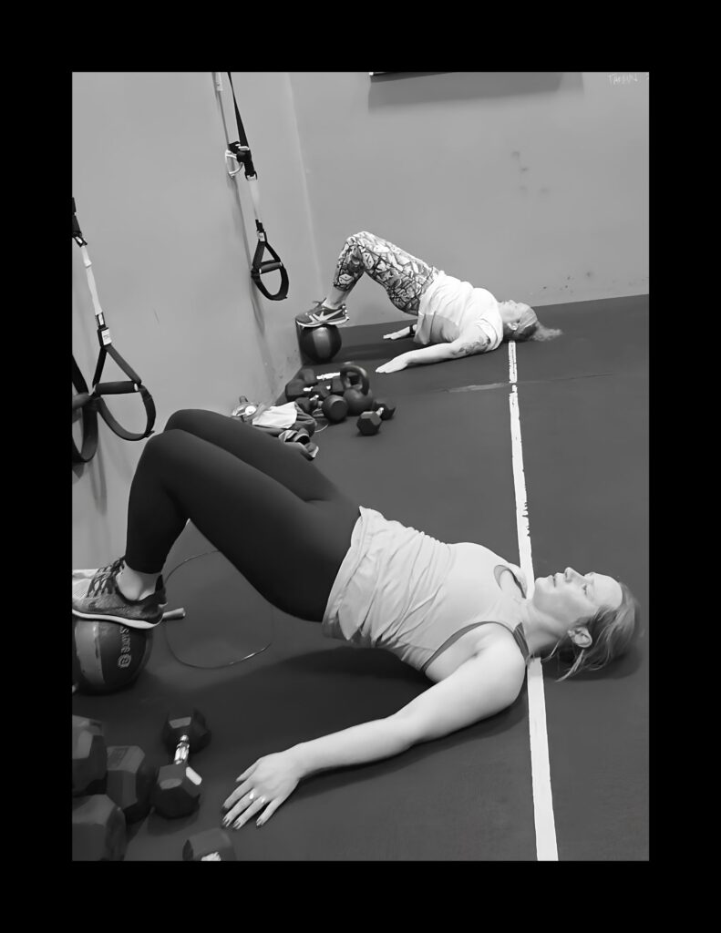 Person lying on a mat in a gym, feet on a wall, performing a glute bridge with a ball and various weights nearby (TRX straps hanging on wall).
