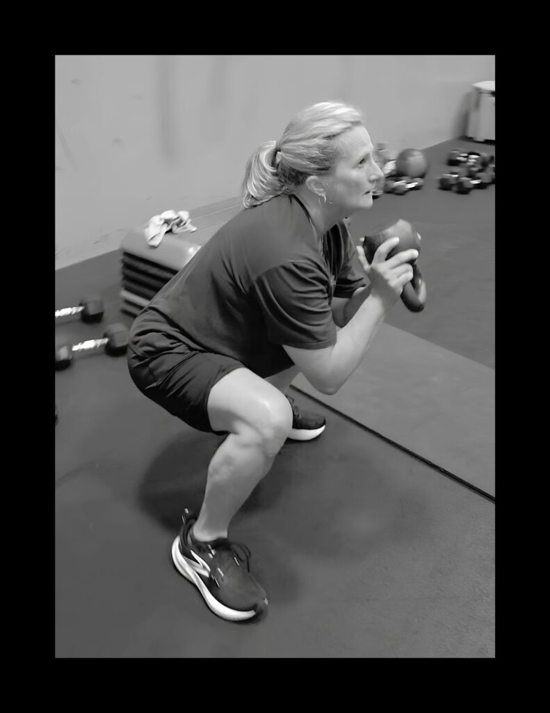 Woman performing a goblet squat in a gym, holding a kettlebell near her chest (black and white).