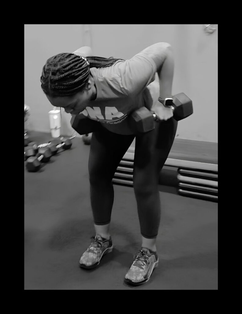 Person in a gym bent over performing a dumbbell row with two dumbbells, braided hair and athletic wear visible.