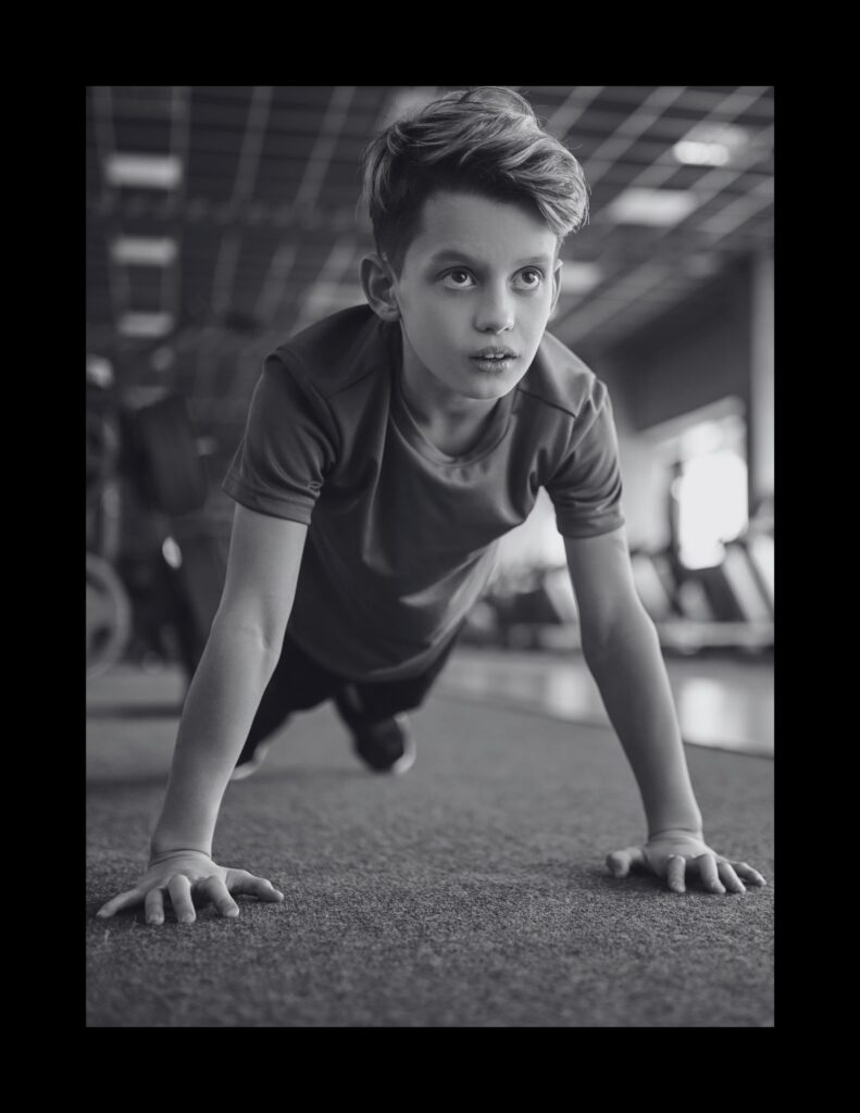 Boy in a gym doing push-ups, hands on the mat, looking upward with a focused expression (black and white photo).