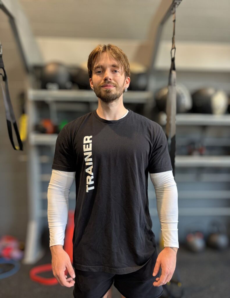 Adult male fitness trainer standing in a gym wearing a black shirt with 'TRAINER' text, white long-sleeve undershirt, neutral smile.